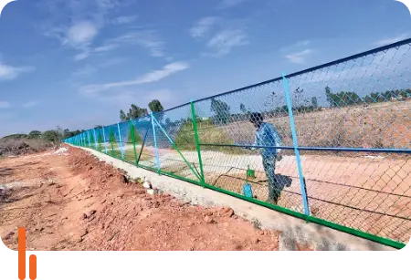 A photograph showing a fence installed to prevent unauthorized access in Devanahalli town, Bangalore Rural district, Karnataka. The image features a sturdy wire fence with a person walking beside it, highlighting efforts to secure the area