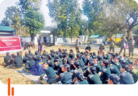 A group of NCC candidates seated outdoors in Dehradun, Uttarakhand, attending a financial literacy and awareness program conducted by ICICI Foundation, with a speaker addressing the audience and military personnel standing nearby.