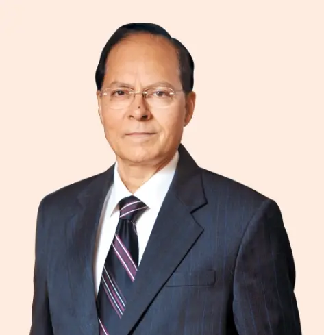 A professional portrait of Chairman wearing a dark suit, tie, and glasses, posing against a light peach background