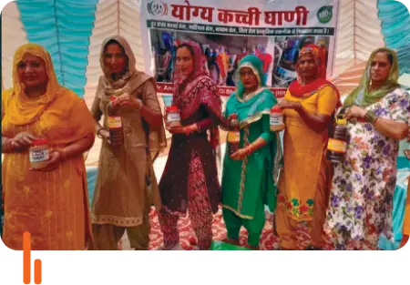 A group of women displaying bottles of mustard oil made by them during a buyer-seller meet at Bharukhera village in Sirsa district, Haryana, with a banner in the background.