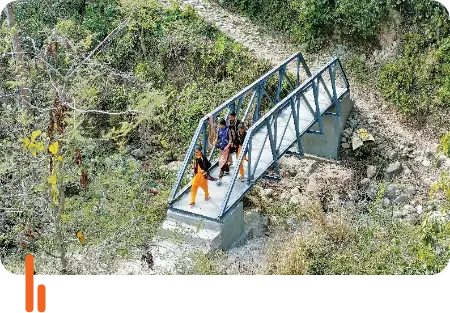 A pedestrian footbridge in a rural area of Jammu and Kashmir, with three people walking across it. The bridge is surrounded by dense foliage and connects two sides of a hilly terrain, providing safe passage for the local community.