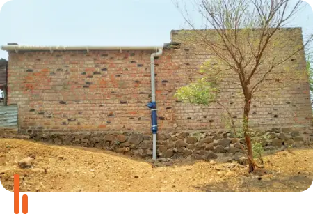 A photograph showing a rainwater harvesting pipe installed on the side of a brick building in Babhalgaon village, Dharashiv district, Maharashtra. The image highlights the sustainable water management practices being implemented in rural areas.