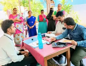 ICICI Bank representatives engaging with a group of local women during a community outreach meeting. The women, dressed in traditional attire, are standing around a table where two bank employees are discussing and reviewing documents. The scene reflects ICICI Bank's efforts in financial inclusion and community support, focusing on empowering local communities through direct engagement and consultation.